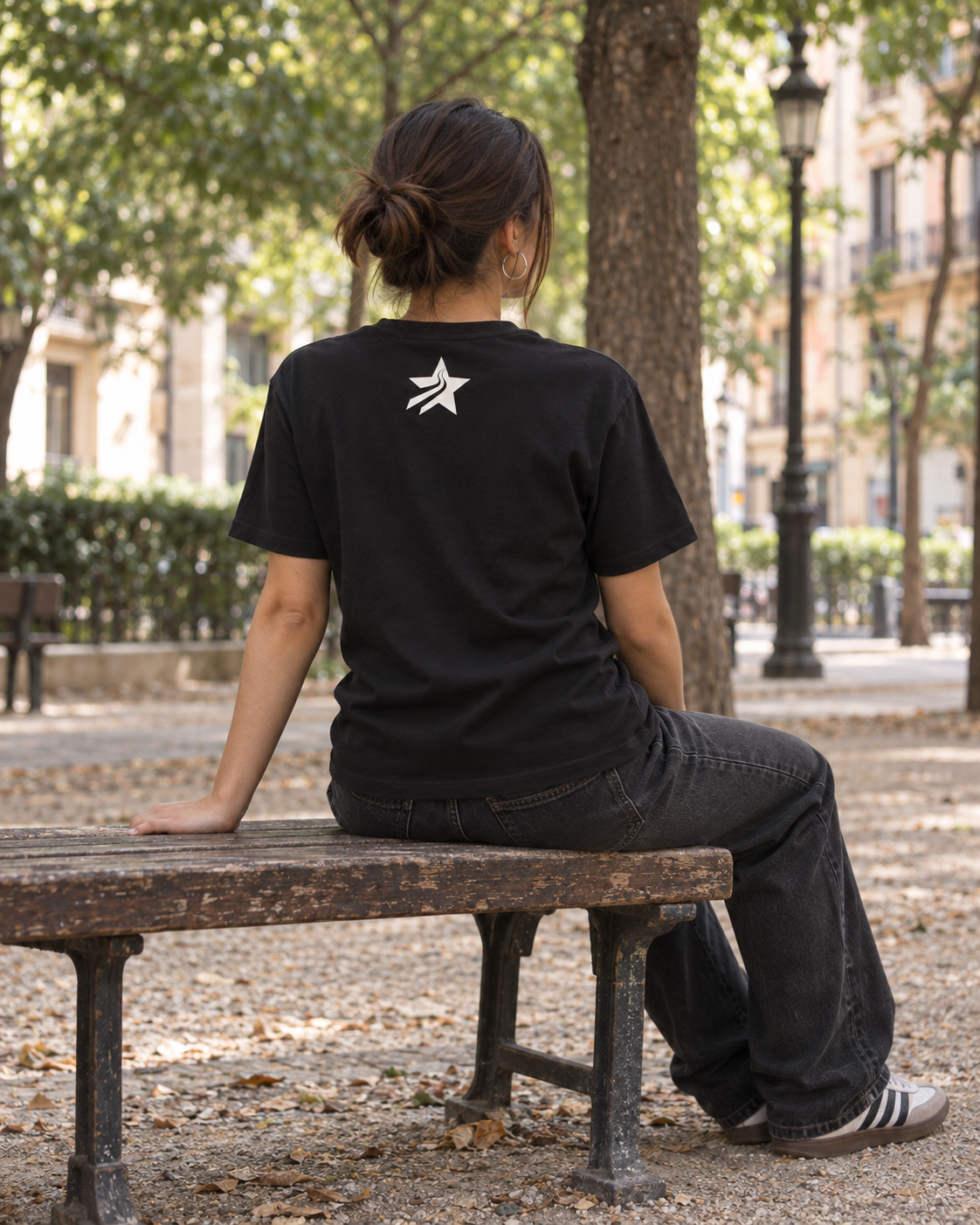 Chica de espaldas sentada en un banco con camiseta negra RG Warrior Lifestyle y estrella blanca bajo el cuello